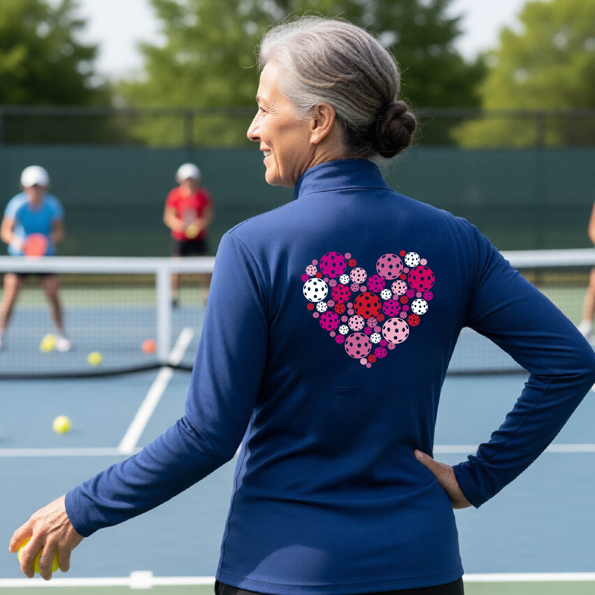 A woman laughing on an outdoor pickleball court wearing a royal blue quarter-zip shirt with a heart-shaped graphic made of pink, red, and white pickleballs on the back. 