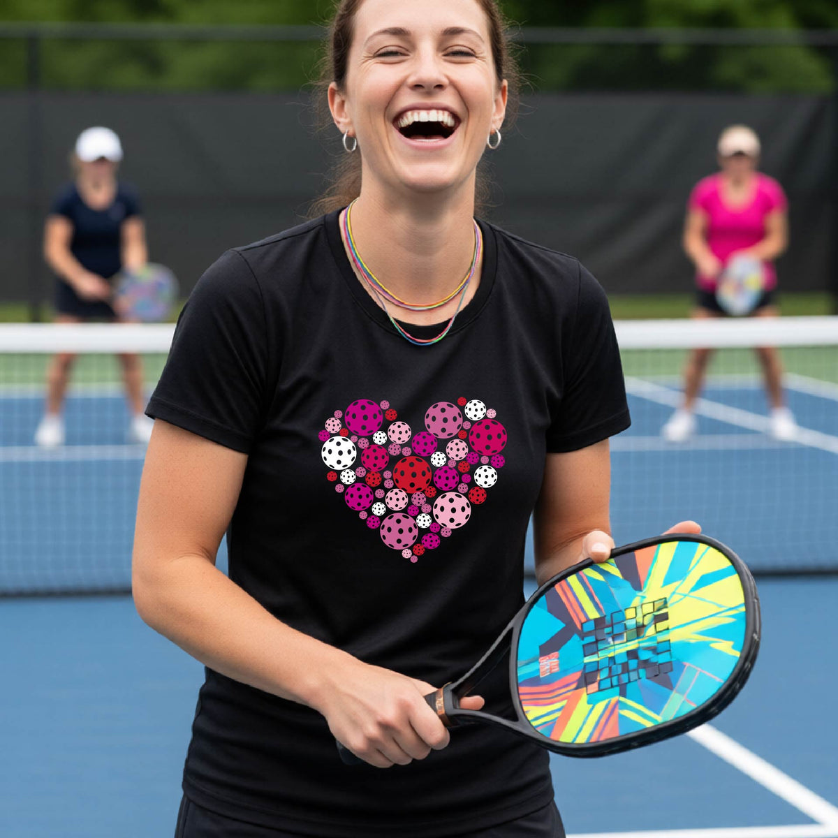 A woman on a pickleball court wearing a black t-shirt with a colorful heart-shaped pattern made of pickleballs. 
