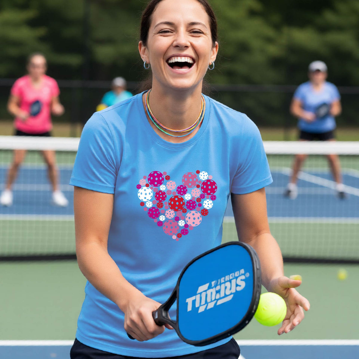 A woman on a pickleball court wearing a blue t-shirt with a colorful heart-shaped pattern made of pickleballs. 