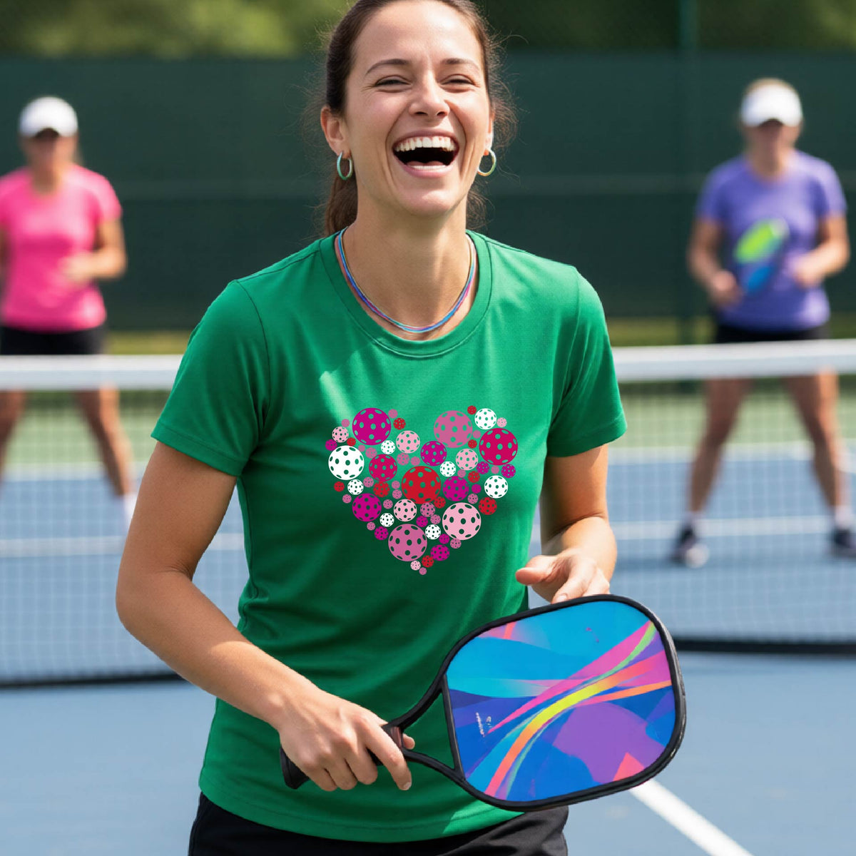 A woman on a pickleball court wearing a green t-shirt with a colorful heart-shaped pattern made of pickleballs. 