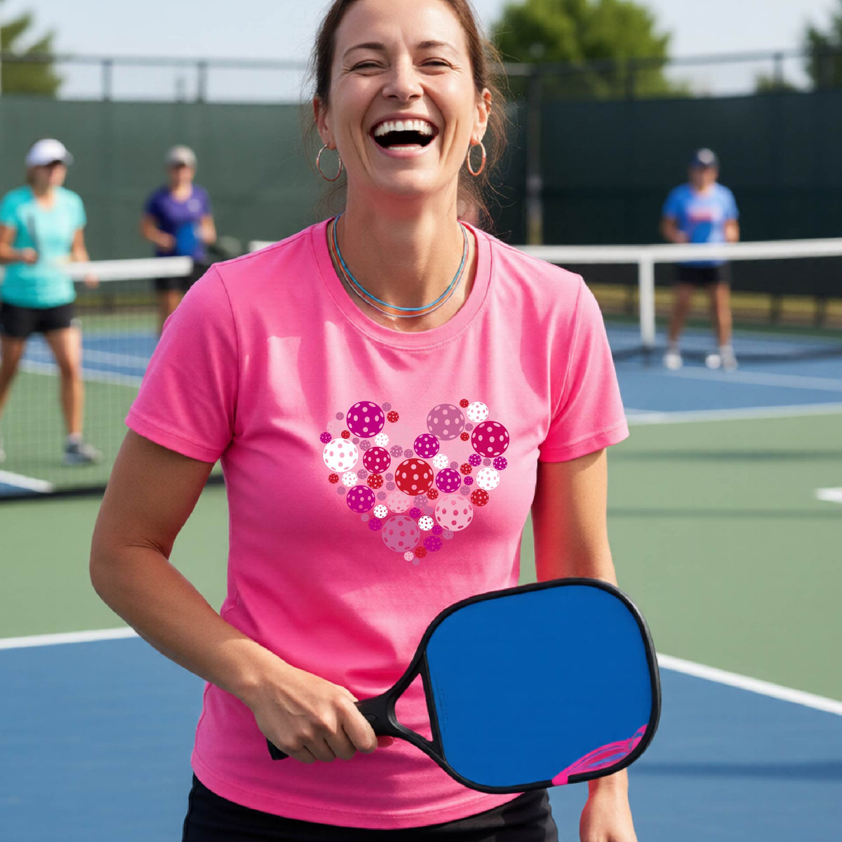 A woman on a pickleball court wearing a pink t-shirt with a colorful heart-shaped pattern made of pickleballs. 