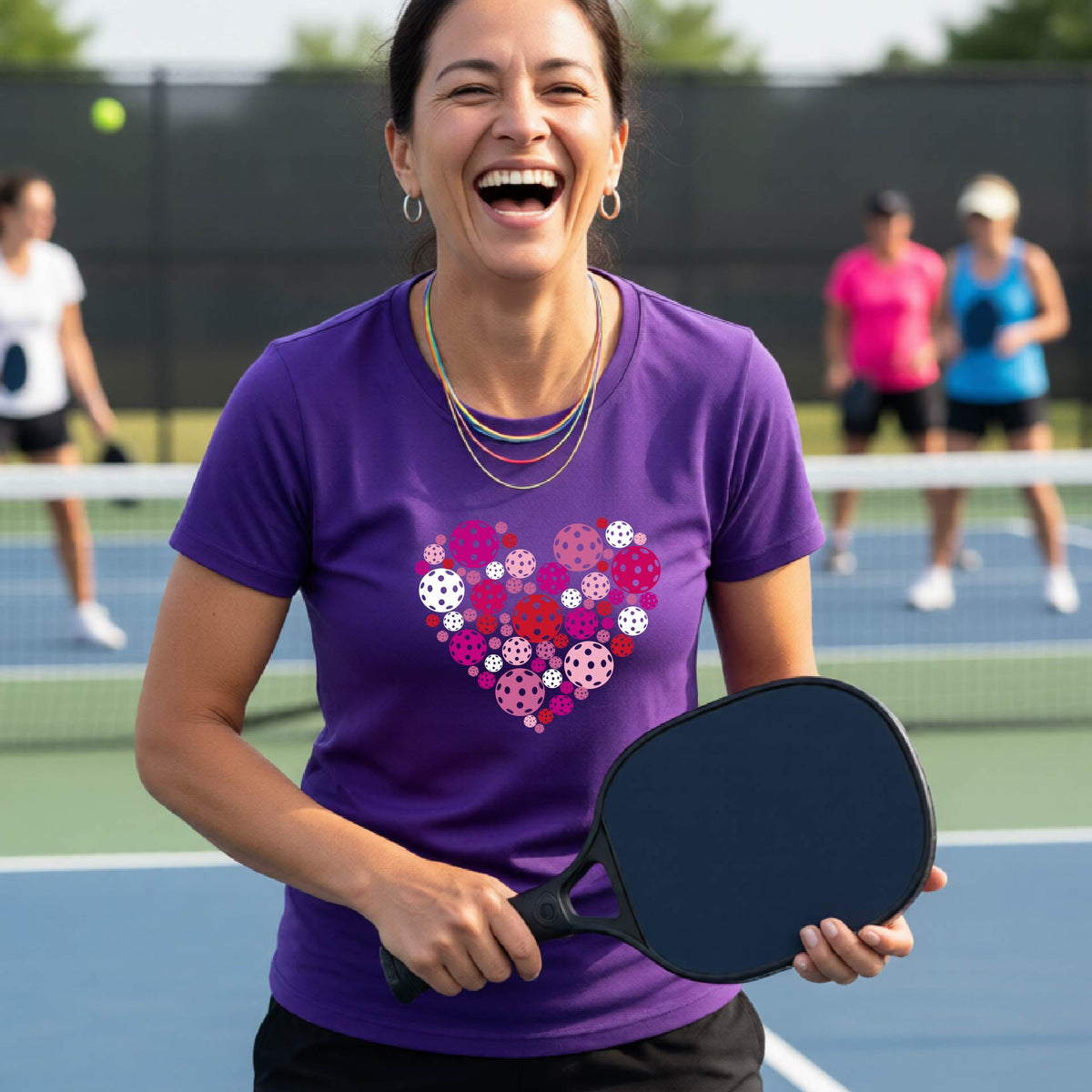 A woman on a pickleball court wearing a purple t-shirt with a colorful heart-shaped pattern made of pickleballs. 