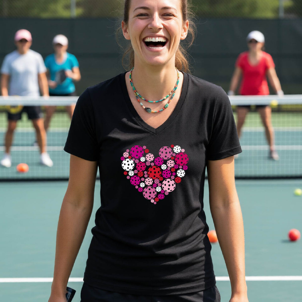A woman smiling on a pickleball court wearing a black v-neck t-shirt with a heart-shaped pattern made of colorful pickleballs on the chest.