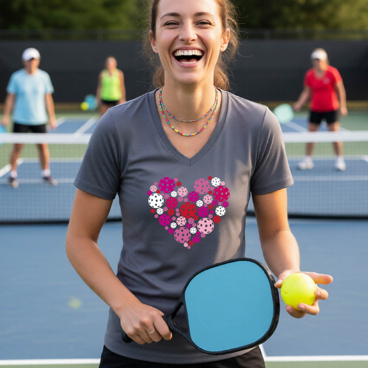 A woman smiling on a pickleball court wearing a gray v-neck t-shirt with a heart-shaped pattern made of colorful pickleballs on the chest.