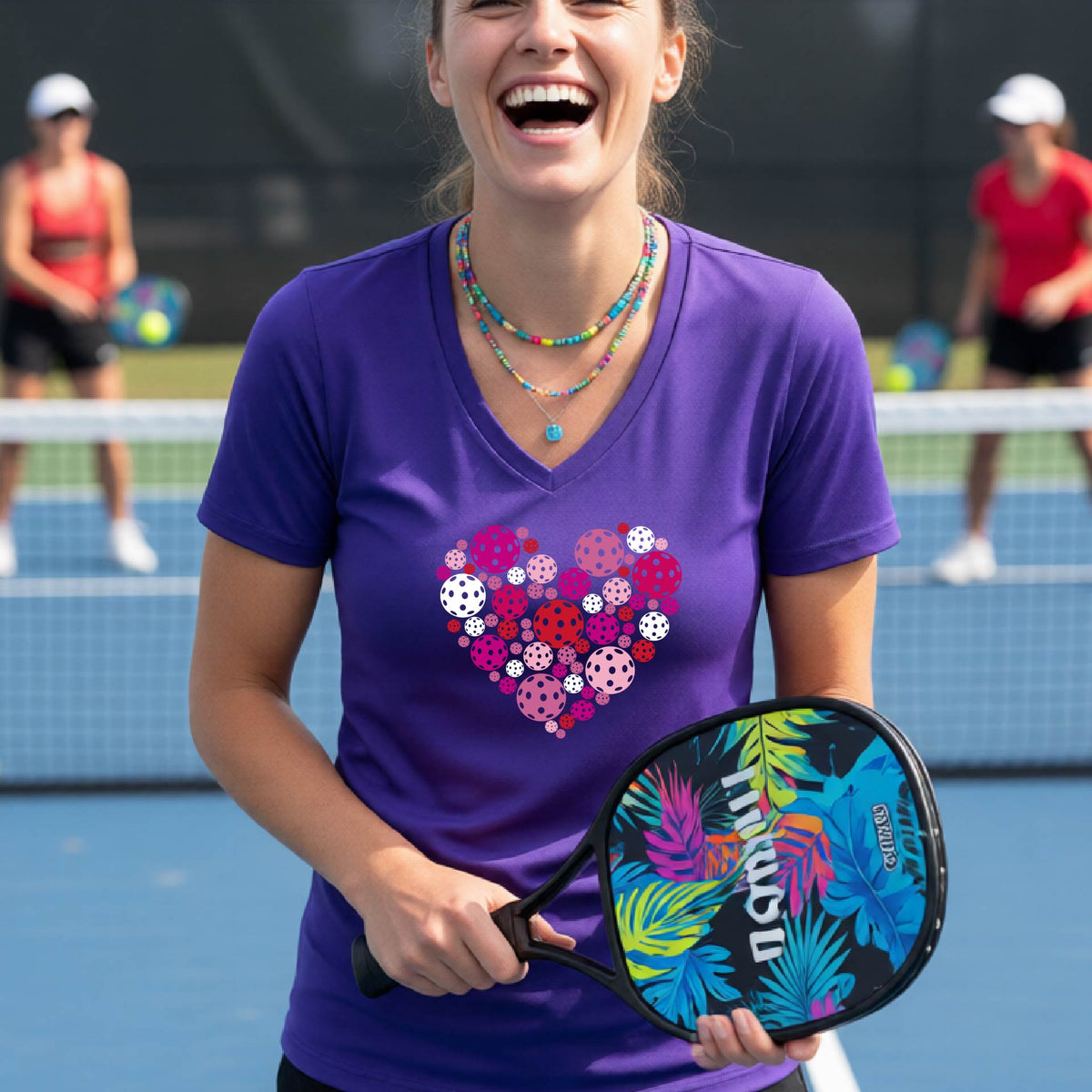 A woman smiling on a pickleball court wearing a purple v-neck t-shirt with a heart-shaped pattern made of colorful pickleballs on the chest.