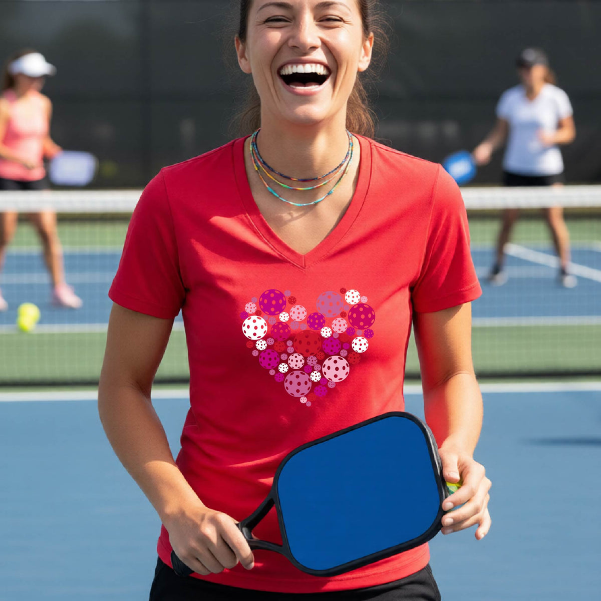 A woman smiling on a pickleball court wearing a red v-neck t-shirt with a heart-shaped pattern made of colorful pickleballs on the chest.