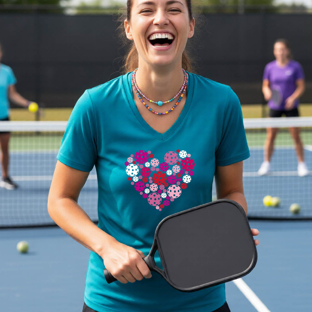 A woman laughing on a pickleball court wearing a teal V-neck t-shirt with a colorful heart-shaped pickelball pattern on the chest.