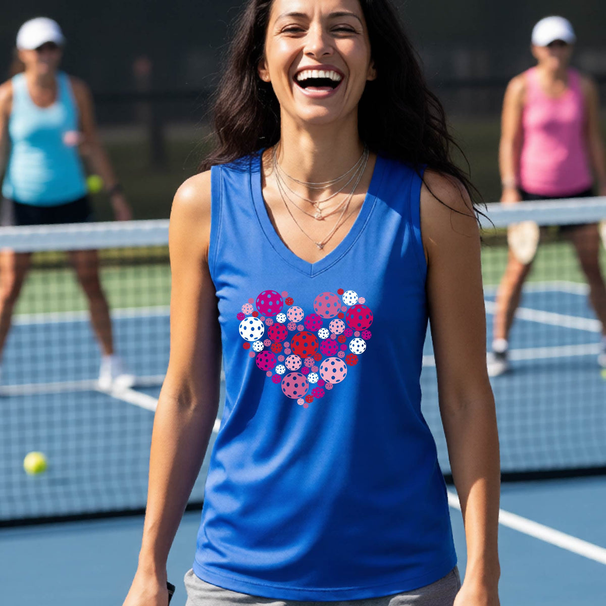 A woman on a sunny outdoor pickleball court wearing a royal blue tank top with a heart-shaped pattern in pink, red, and purple pickleballs. 