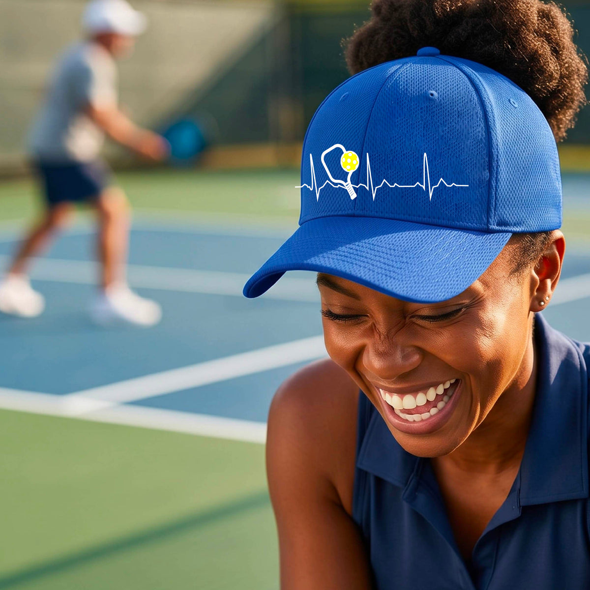 A close-up of a royal blue athletic hat showing a white pickleball graphic of a heartbeat EKG including a pickleball paddle and a yellow pickleball, in front of a pickleball court.