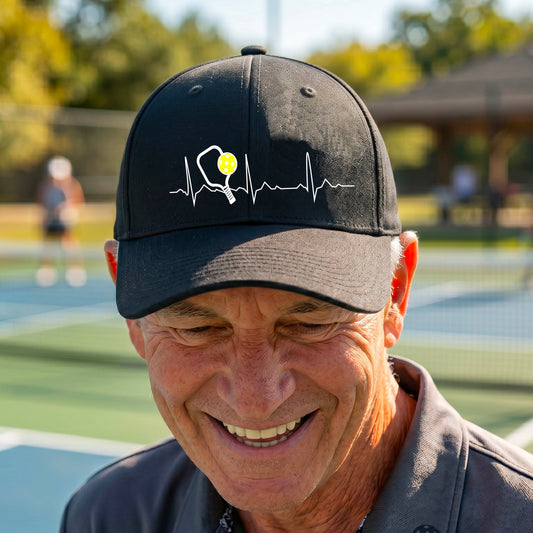 A close-up of a black athletic hat showing a white pickleball graphic of a heartbeat EKG including a pickleball paddle and a yellow pickleball, in front of a pickleball court.