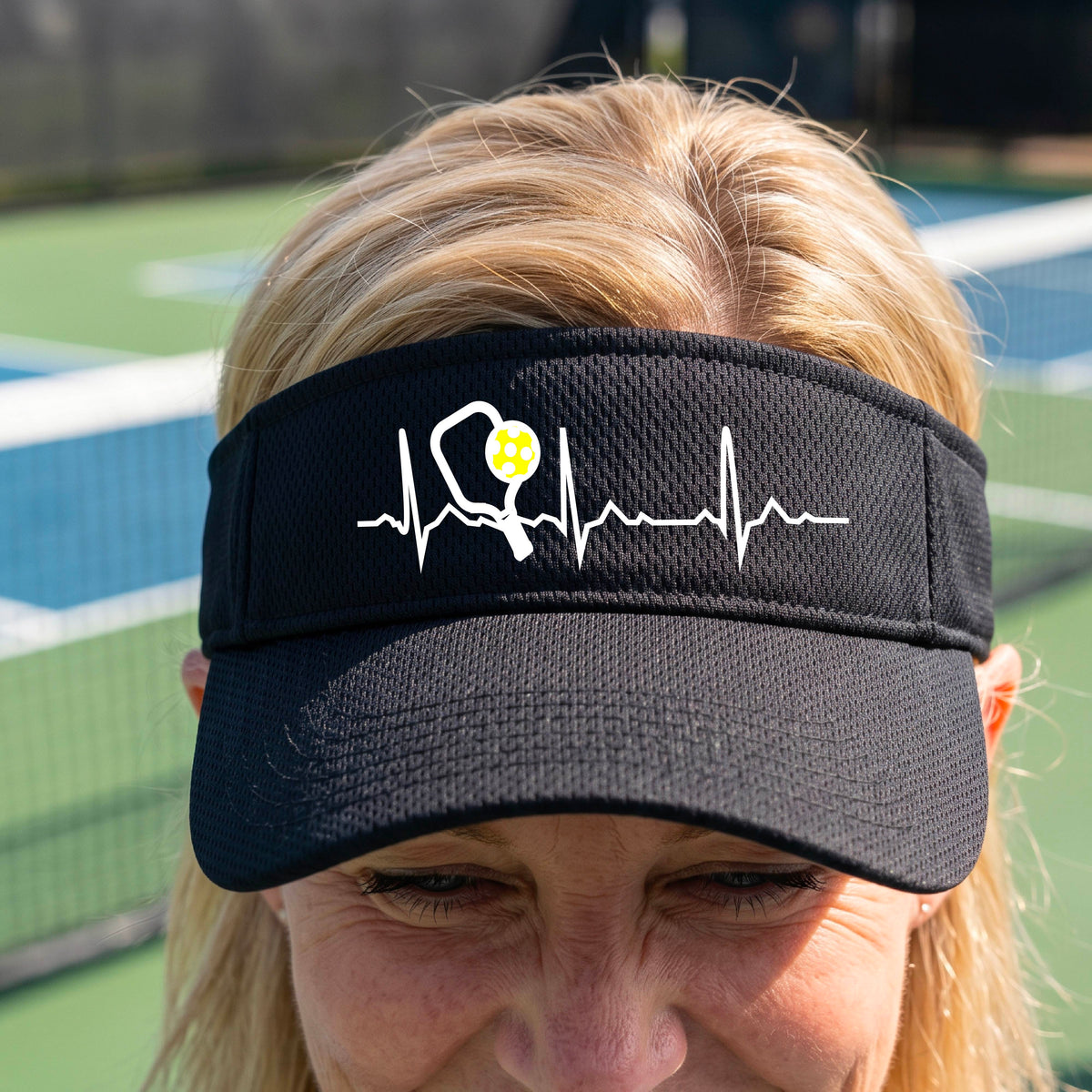 A close-up of a black athletic visor showing a white pickleball graphic of a heartbeat EKG including a pickleball paddle and a yellow pickleball, in front of a pickleball court.