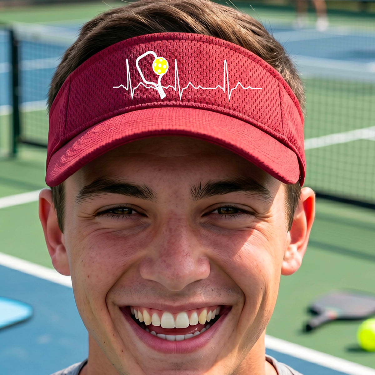 A close-up of a red athletic visor showing a white pickleball graphic of a heartbeat EKG including a pickleball paddle and a yellow pickleball, in front of a pickleball court.