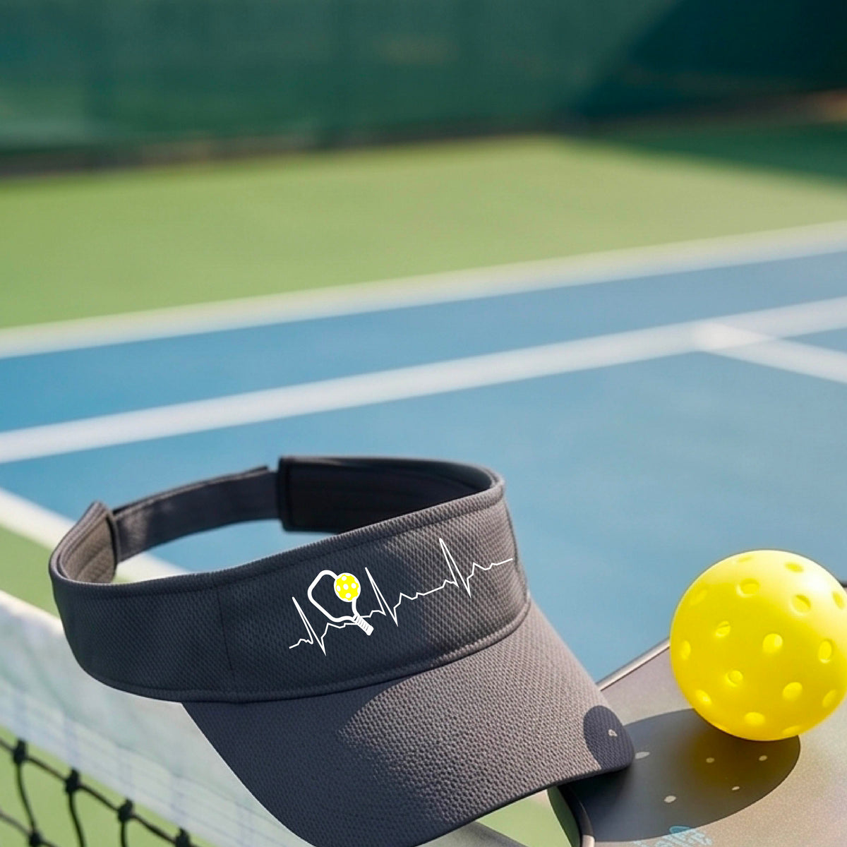 A close-up of a gray athletic visor showing a white pickleball graphic of a heartbeat EKG including a pickleball paddle and a yellow pickleball, in front of a pickleball court.