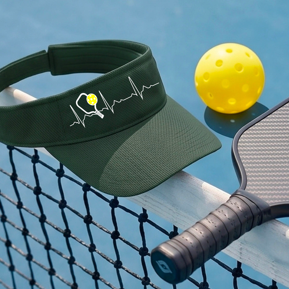 A close-up of a green athletic visor showing a white pickleball graphic of a heartbeat EKG including a pickleball paddle and a yellow pickleball, in front of a pickleball court.