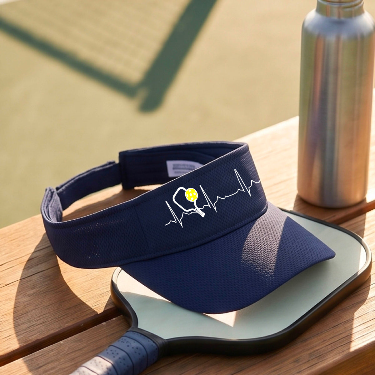 A close-up of a navy blue athletic visor showing a white pickleball graphic of a heartbeat EKG including a pickleball paddle and a yellow pickleball, in front of a pickleball court.