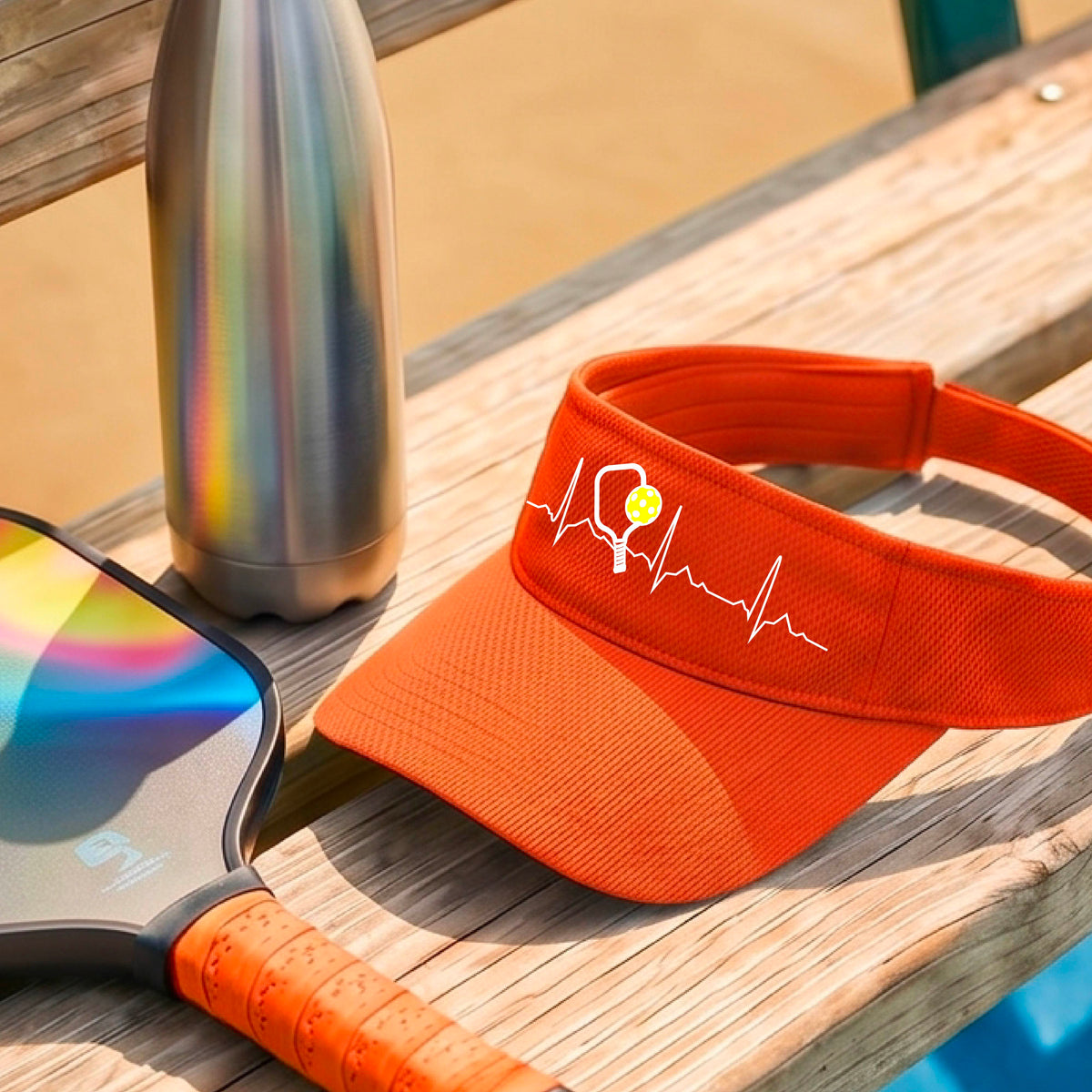 A close-up of an orange athletic visor showing a white pickleball graphic of a heartbeat EKG including a pickleball paddle and a yellow pickleball, in front of a pickleball court.