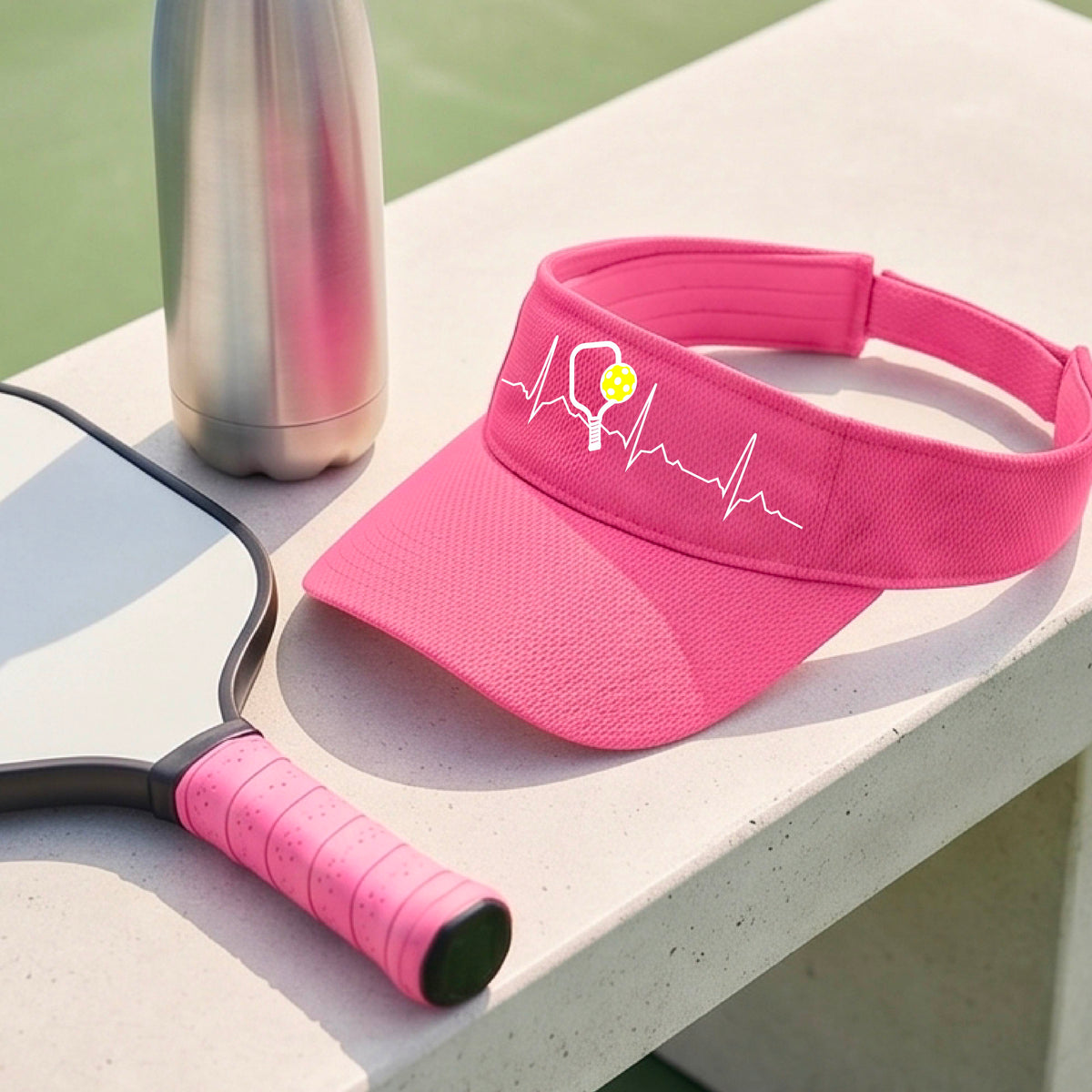 A close-up of a pink athletic visor showing a white pickleball graphic of a heartbeat EKG including a pickleball paddle and a yellow pickleball, in front of a pickleball court.