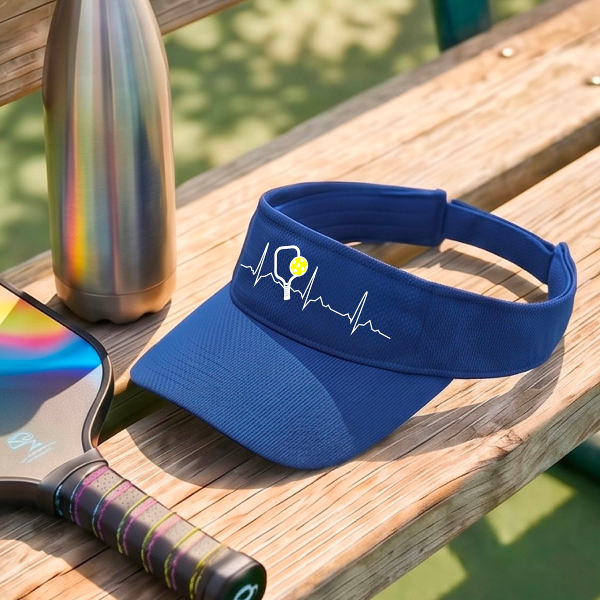A close-up of a royal blue athletic visor showing a white pickleball graphic of a heartbeat EKG including a pickleball paddle and a yellow pickleball, in front of a pickleball court.