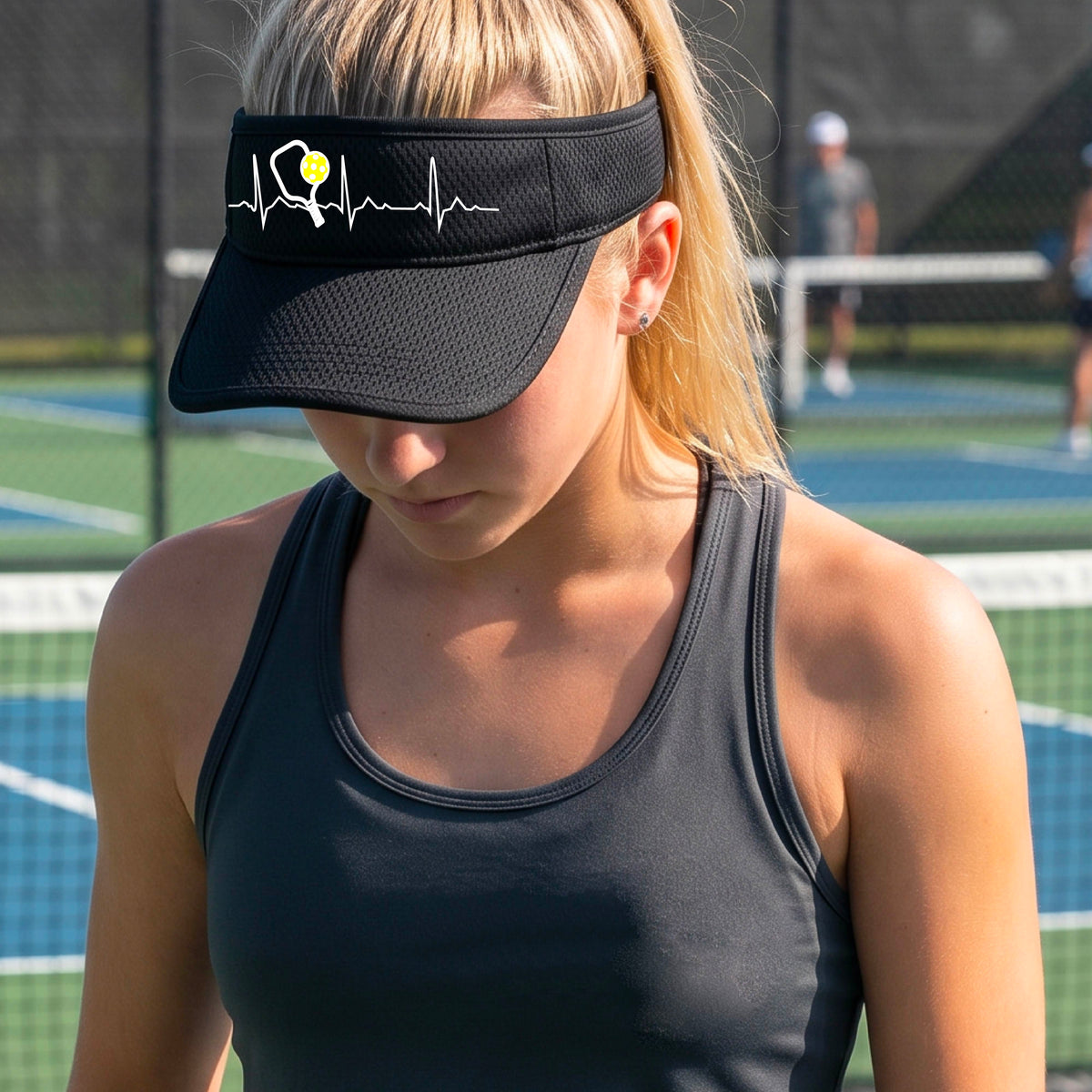 A close-up of a black athletic visor showing a white pickleball graphic of a heartbeat EKG including a pickleball paddle and a yellow pickleball, in front of a pickleball court.