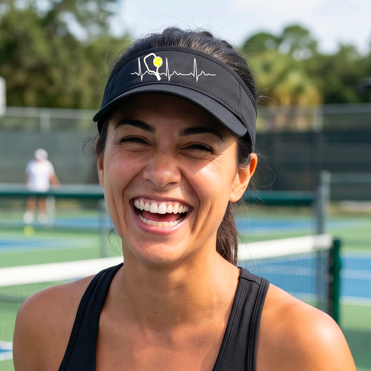A close-up of a black athletic visor showing a white pickleball graphic of a heartbeat EKG including a pickleball paddle and a yellow pickleball, in front of a pickleball court.