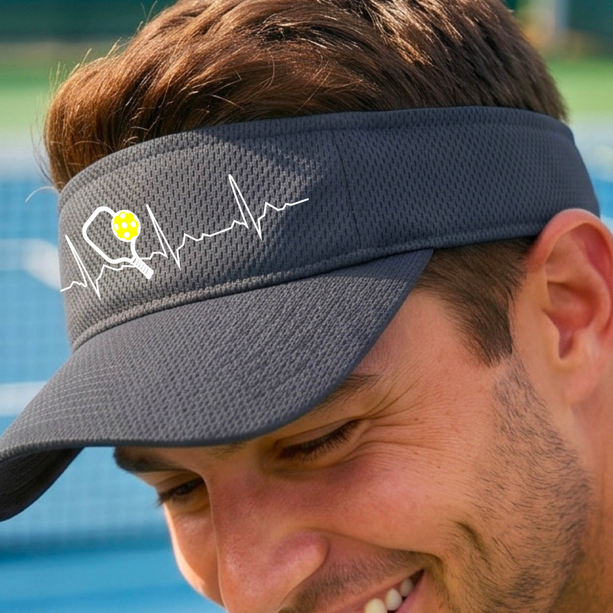 A close-up of a gray athletic visor showing a white pickleball graphic of a heartbeat EKG including a pickleball paddle and a yellow pickleball, in front of a pickleball court.
