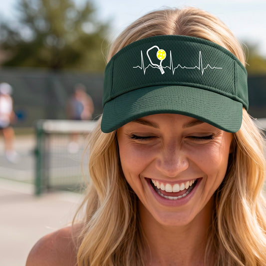 A close-up of a green athletic visor showing a white pickleball graphic of a heartbeat EKG including a pickleball paddle and a yellow pickleball, in front of a pickleball court.