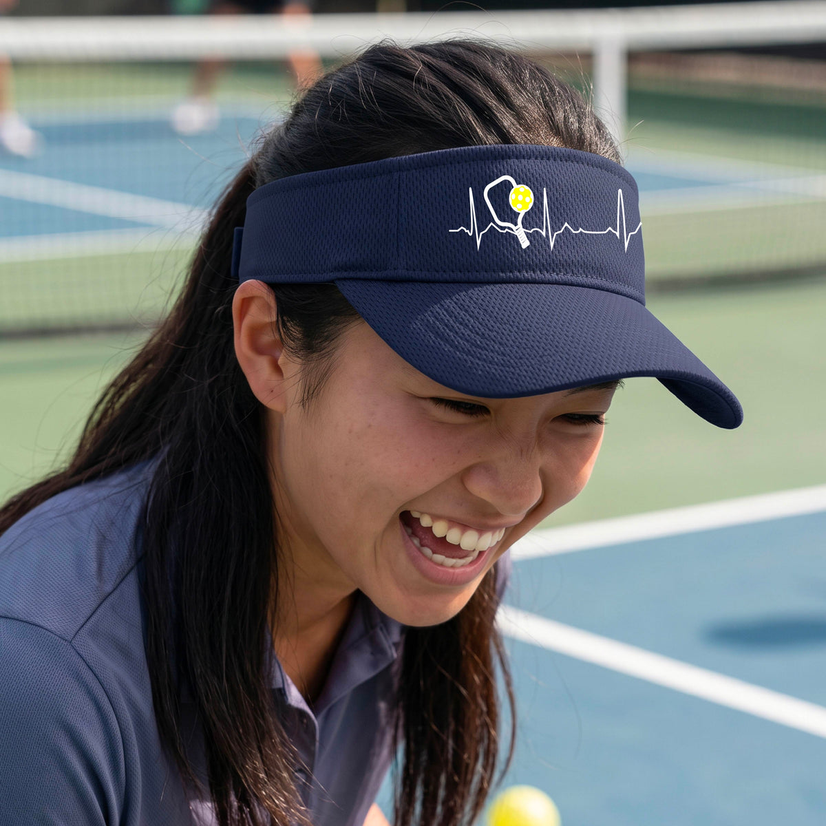 A close-up of a navy blue athletic visor showing a white pickleball graphic of a heartbeat EKG including a pickleball paddle and a yellow pickleball, in front of a pickleball court.