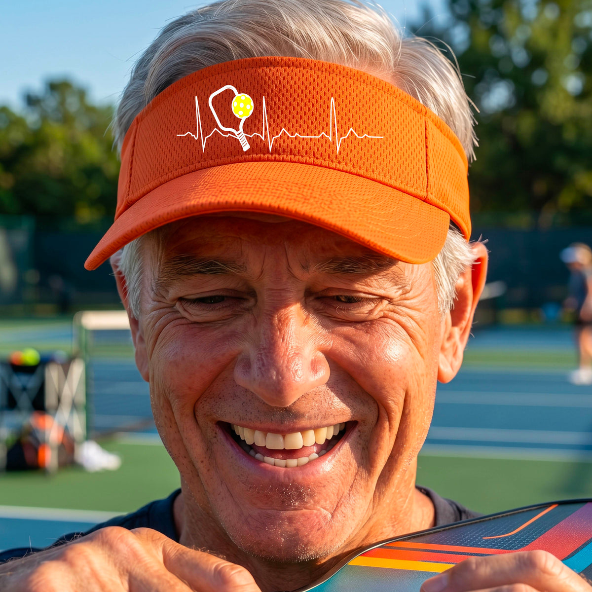 A close-up of an orange athletic visor showing a white pickleball graphic of a heartbeat EKG including a pickleball paddle and a yellow pickleball, in front of a pickleball court.