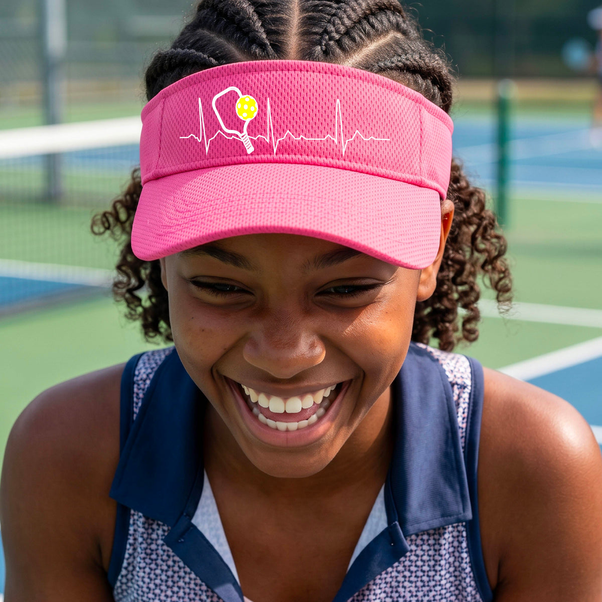 A close-up of a pink athletic visor showing a white pickleball graphic of a heartbeat EKG including a pickleball paddle and a yellow pickleball, in front of a pickleball court.
