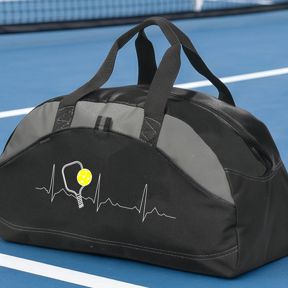 A close-up of a black and gray medium size duffel bag with a white pickleball graphic of a heartbeat EKG including a pickleball paddle and a yellow pickleball, in front of a pickleball court.