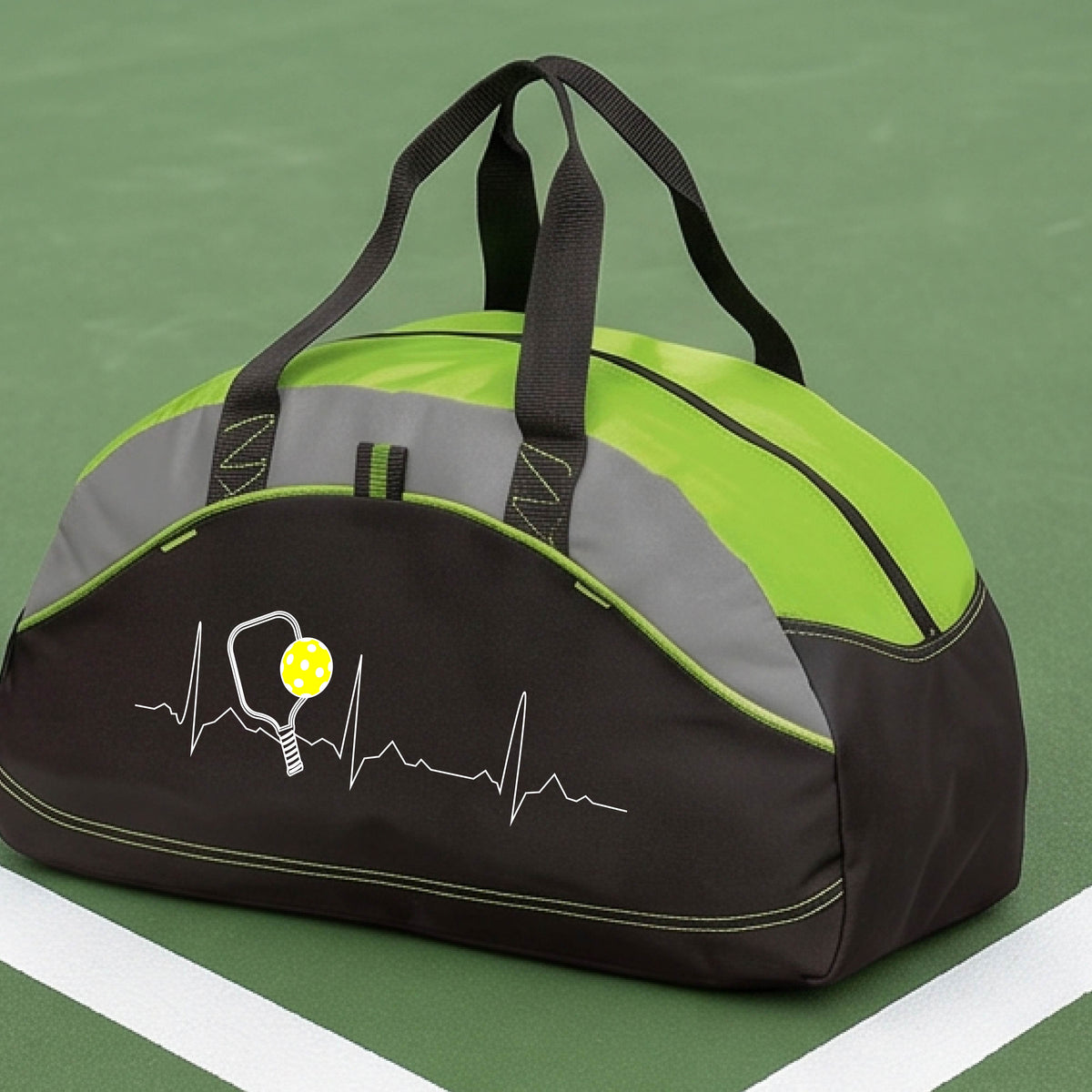 A close-up of a lime green, black and gray medium size duffel bag with a white pickleball graphic of a heartbeat EKG including a pickleball paddle and a yellow pickleball, in front of a pickleball court.