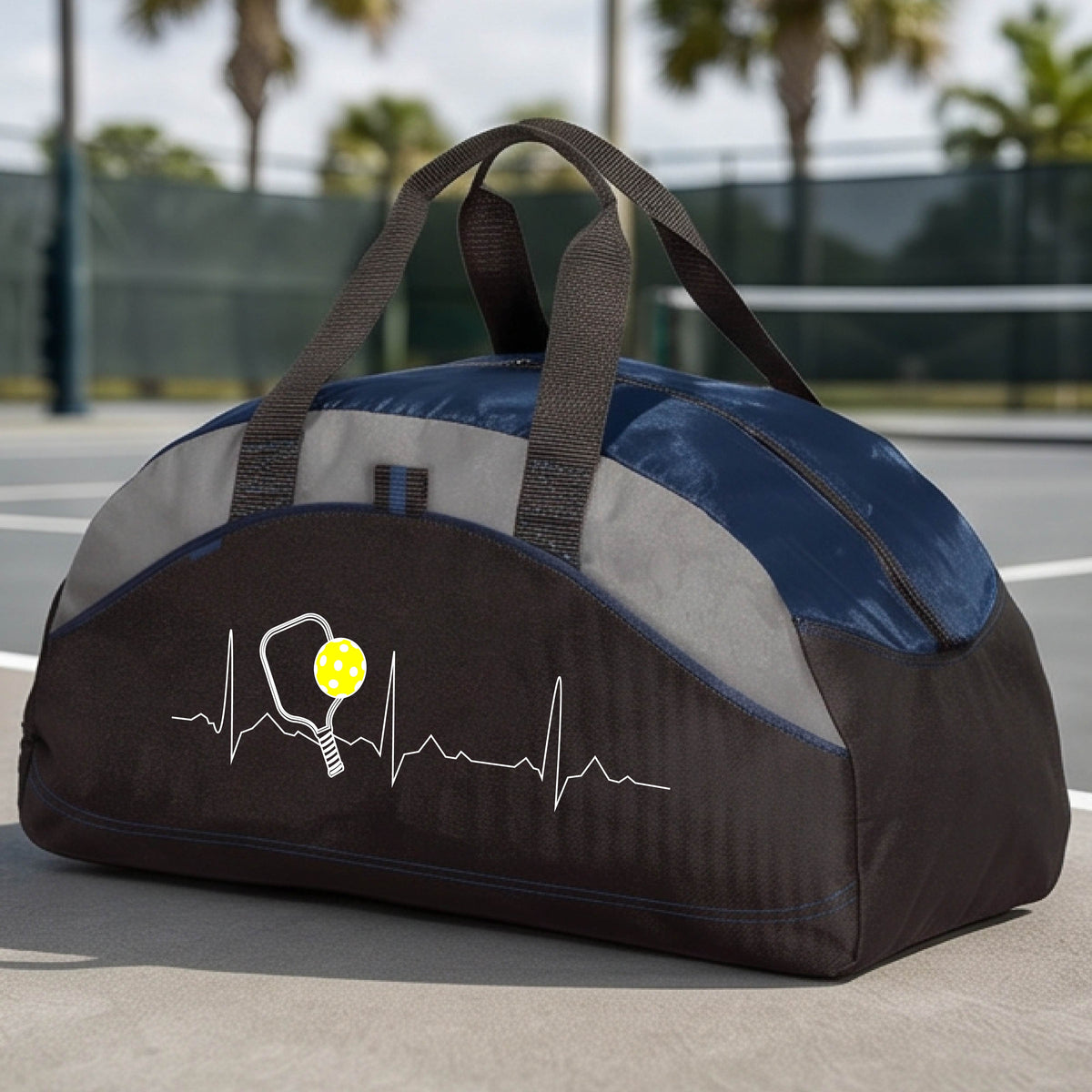 A close-up of a navy blue, black and gray medium size duffel bag with a white pickleball graphic of a heartbeat EKG including a pickleball paddle and a yellow pickleball, in front of a pickleball court.