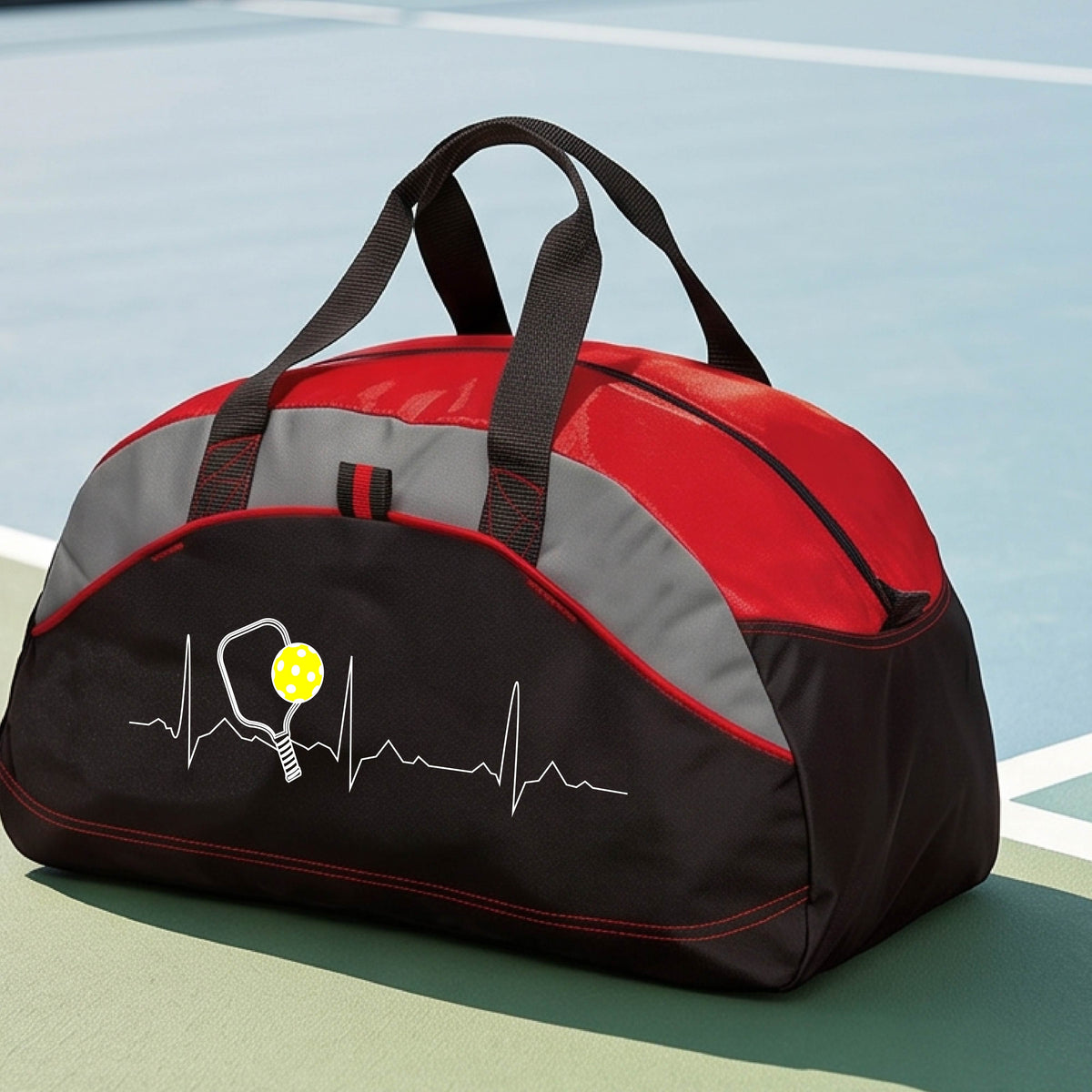 A close-up of a red, black and gray medium size duffel bag with a white pickleball graphic of a heartbeat EKG including a pickleball paddle and a yellow pickleball, in front of a pickleball court.
