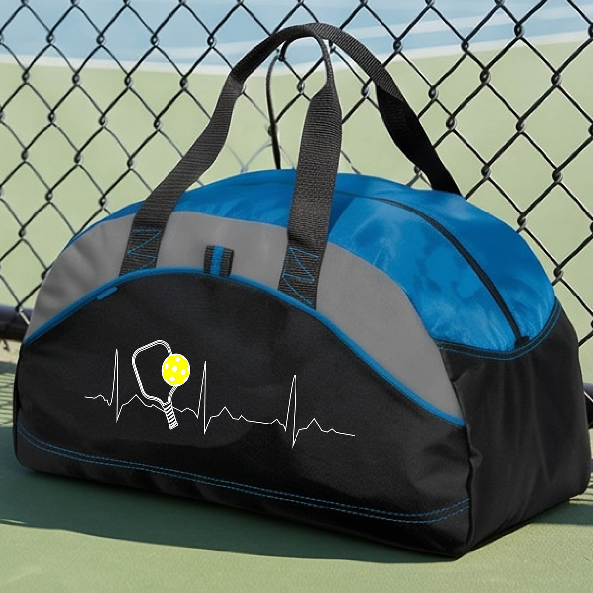 A close-up of a royal blue, black and gray medium size duffel bag with a white pickleball graphic of a heartbeat EKG including a pickleball paddle and a yellow pickleball, in front of a pickleball court.