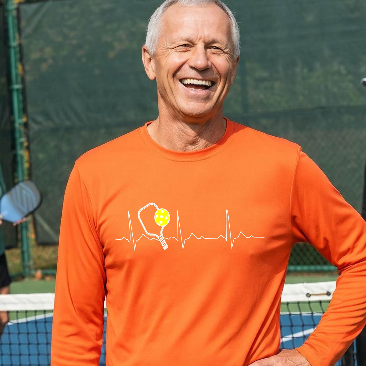 A close-up of an orange mens long-sleeve t-shirt showing a white pickleball graphic of a heartbeat EKG including a pickleball paddle and a yellow pickleball, in front of a pickleball court.
