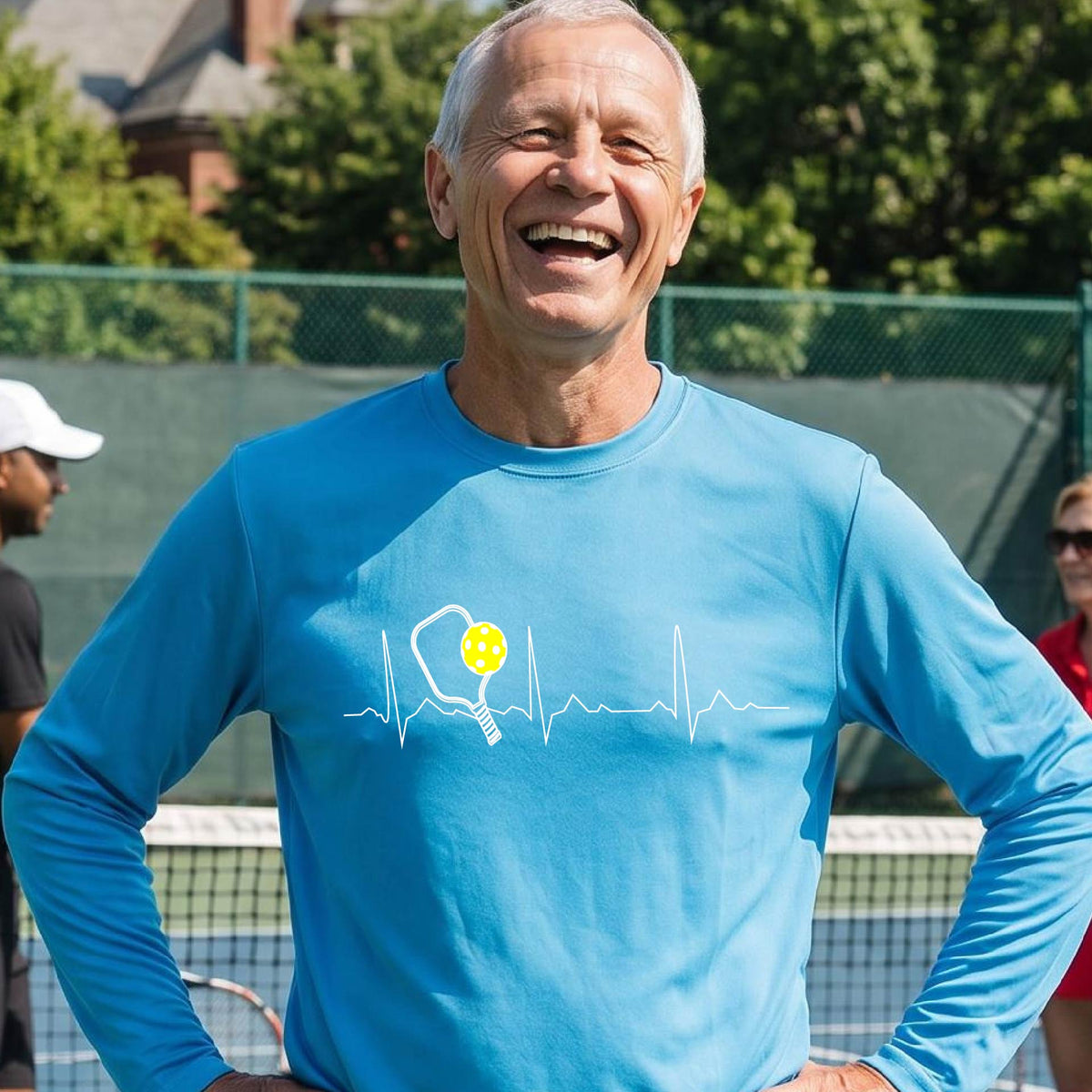A close-up of a bright-blue mens long-sleeve t-shirt showing a white pickleball graphic of a heartbeat EKG including a pickleball paddle and a yellow pickleball, in front of a pickleball court.