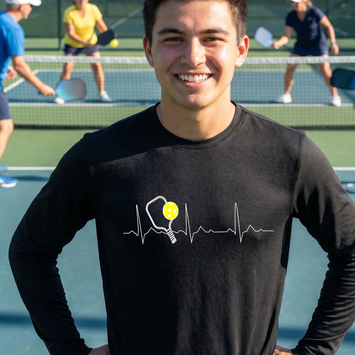 A close-up of a black mens long-sleeve showing a white pickleball graphic of a heartbeat EKG including a pickleball paddle and a yellow pickleball, in front of a pickleball court.