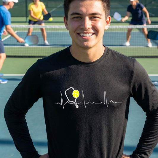 A close-up of a black mens long-sleeve showing a white pickleball graphic of a heartbeat EKG including a pickleball paddle and a yellow pickleball, in front of a pickleball court.