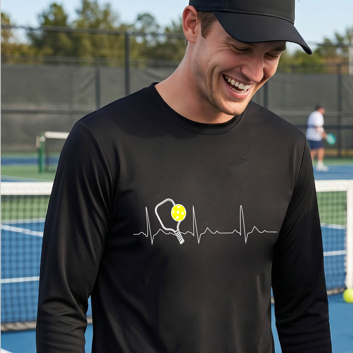 A close-up of a black mens long-sleeve t-shirt showing a white pickleball graphic of a heartbeat EKG including a pickleball paddle and a yellow pickleball, in front of a pickleball court.