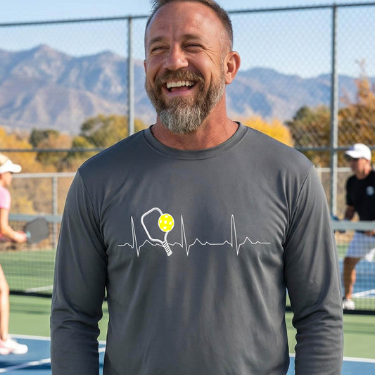 A close-up of a gray mens long-sleeve t-shirt showing a white pickleball graphic of a heartbeat EKG including a pickleball paddle and a yellow pickleball, in front of a pickleball court.