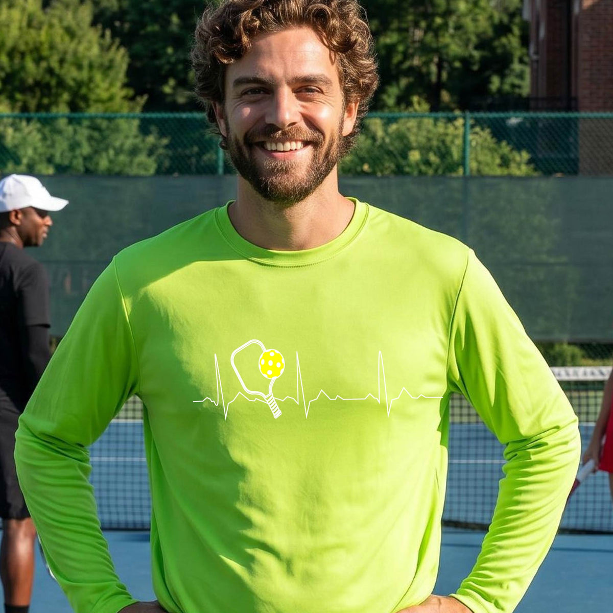 A close-up of a lime green mens long-sleeve t-shirt showing a white pickleball graphic of a heartbeat EKG including a pickleball paddle and a yellow pickleball, in front of a pickleball court.