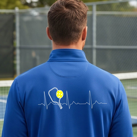 A close-up of a mans royal blue quarter-zip shirt showing a white pickleball graphic of a heartbeat EKG including a pickleball paddle and a yellow pickleball, in front of a pickleball court.