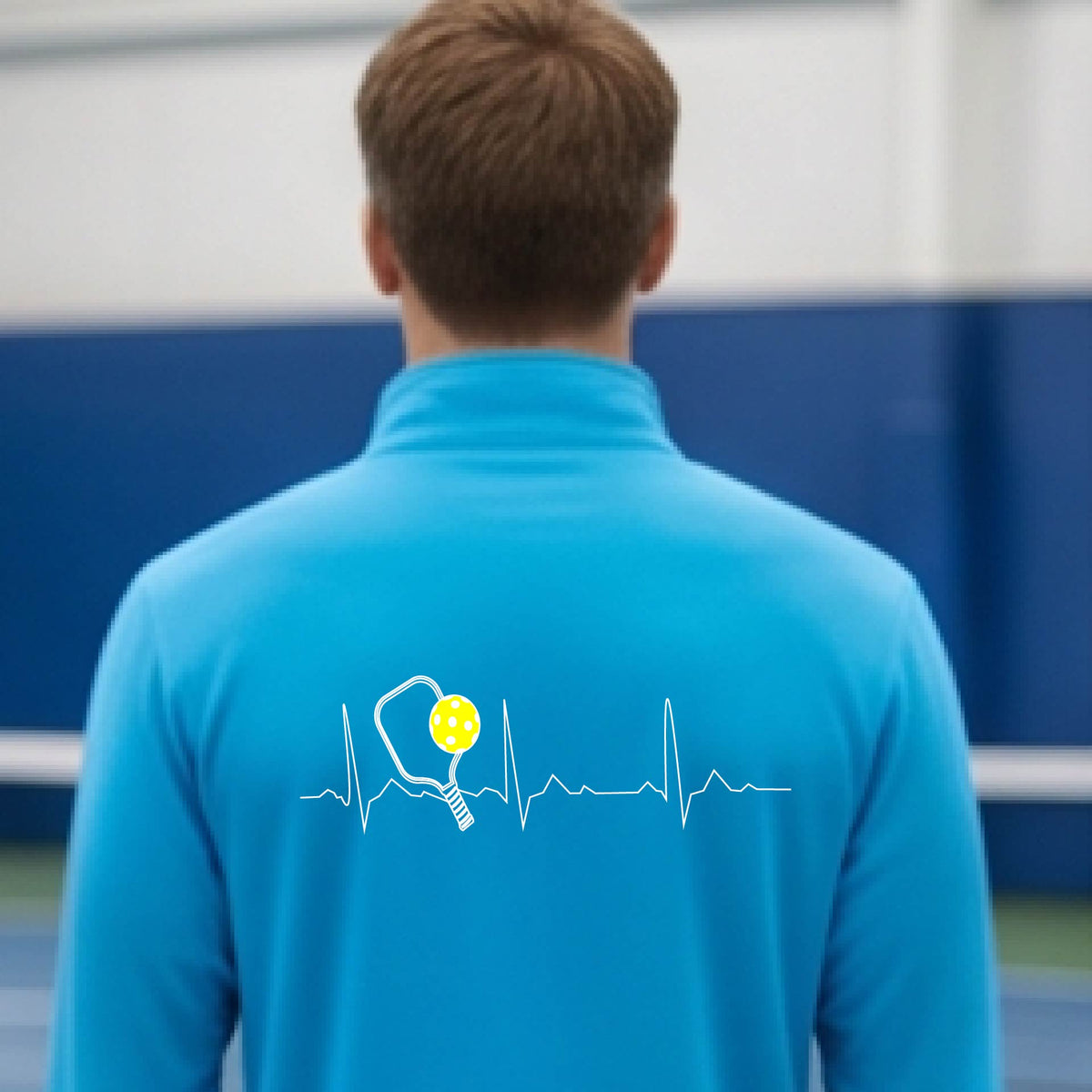A close-up of a mans bright blue quarter-zip shirt showing a white pickleball graphic of a heartbeat EKG including a pickleball paddle and a yellow pickleball, in front of a pickleball court.