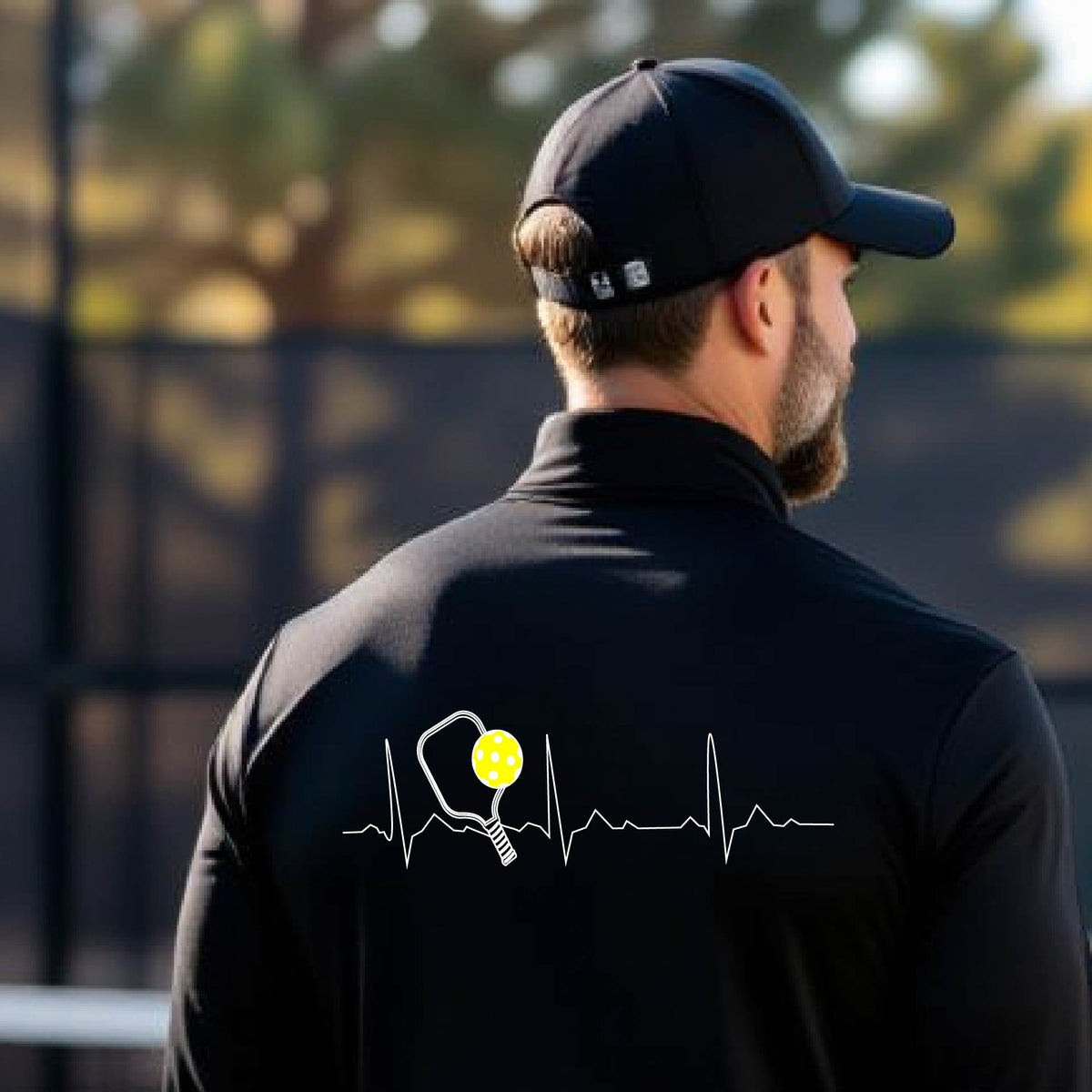 A close-up of a mans black quarter-zip shirt showing a white pickleball graphic of a heartbeat EKG including a pickleball paddle and a yellow pickleball, in front of a pickleball court.