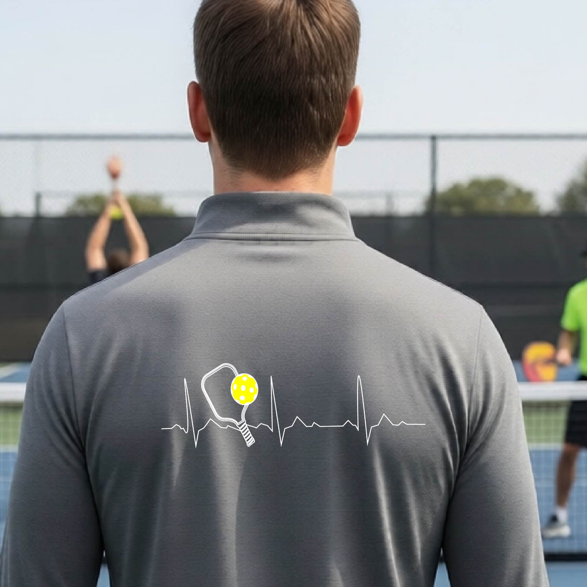 A close-up of a mans gray quarter-zip shirt showing a white pickleball graphic of a heartbeat EKG including a pickleball paddle and a yellow pickleball, in front of a pickleball court.
