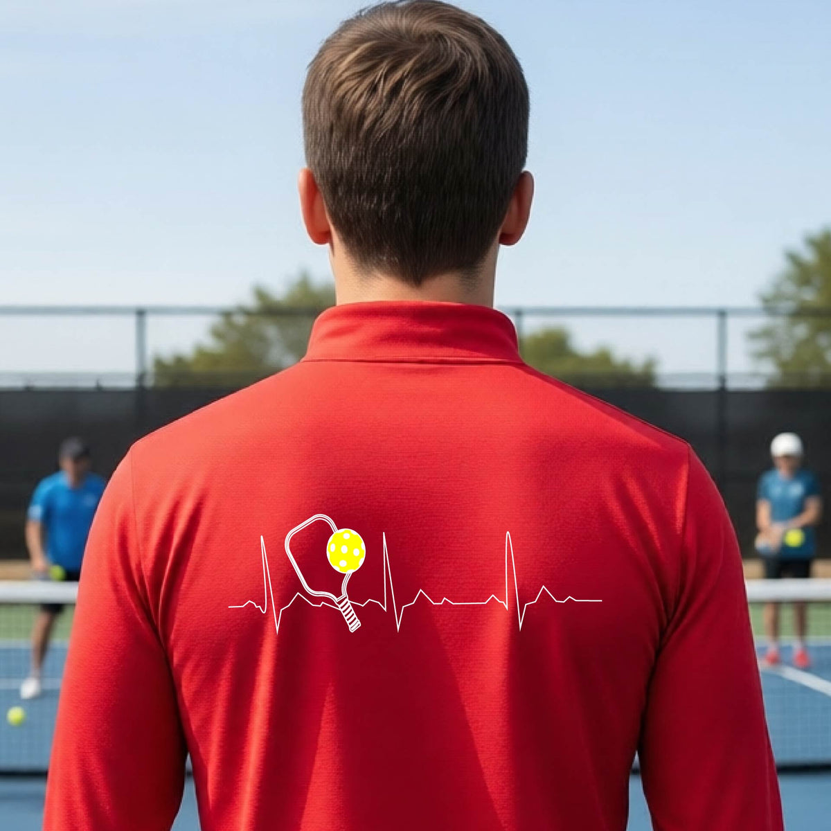 A close-up of a mans red quarter-zip shirt showing a white pickleball graphic of a heartbeat EKG including a pickleball paddle and a yellow pickleball, in front of a pickleball court.