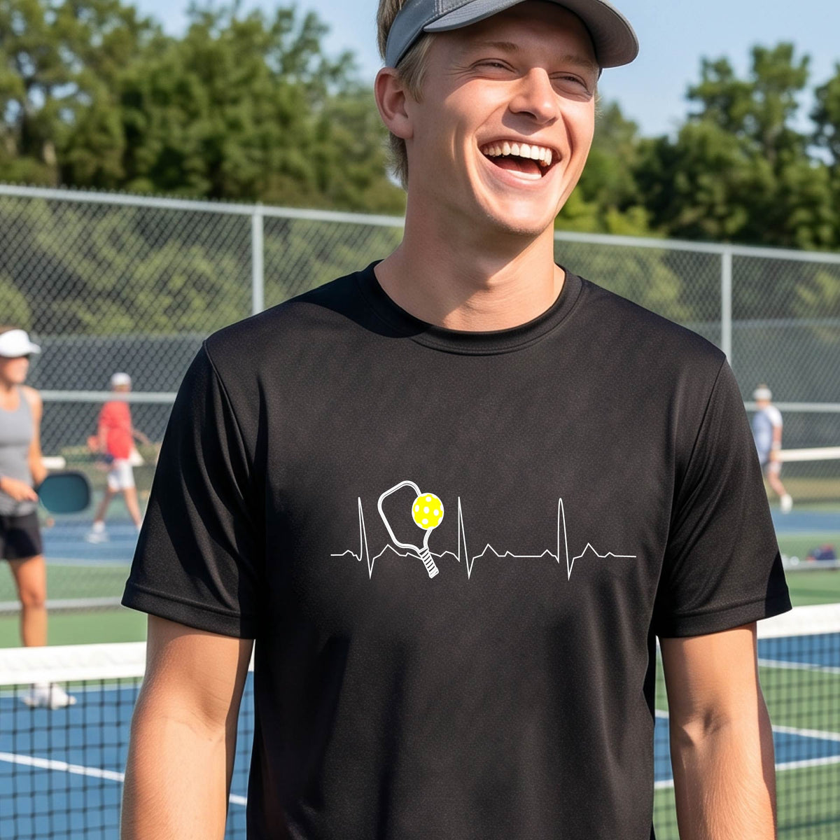 A close-up of a man in a black short-sleeve t-shirt sporting a white pickleball graphic of a heartbeat EKG including a pickleball paddle and a yellow pickleball across the check, in front of a pickleball court.