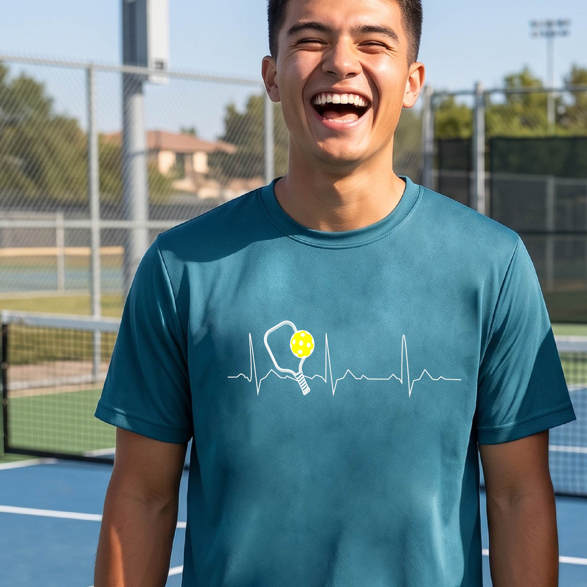A close-up of a man in a teal short-sleeve t-shirt sporting a white pickleball graphic of a heartbeat EKG including a pickleball paddle and a yellow pickleball across the check, in front of a pickleball court.

