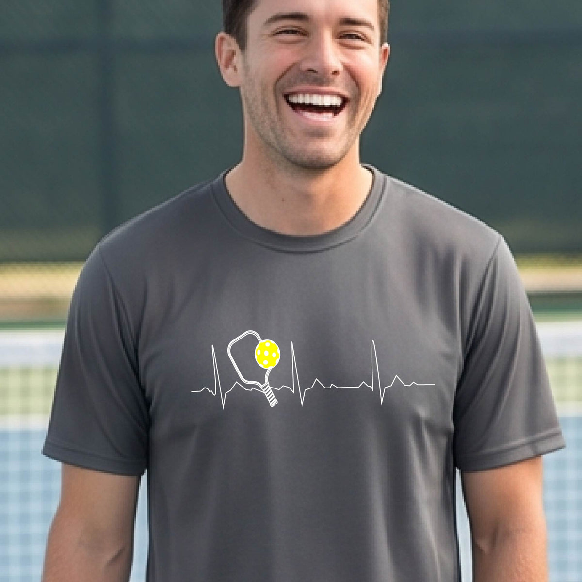 A close-up of a man in a gray short-sleeve t-shirt sporting a white pickleball graphic of a heartbeat EKG including a pickleball paddle and a yellow pickleball across the check, in front of a pickleball court.