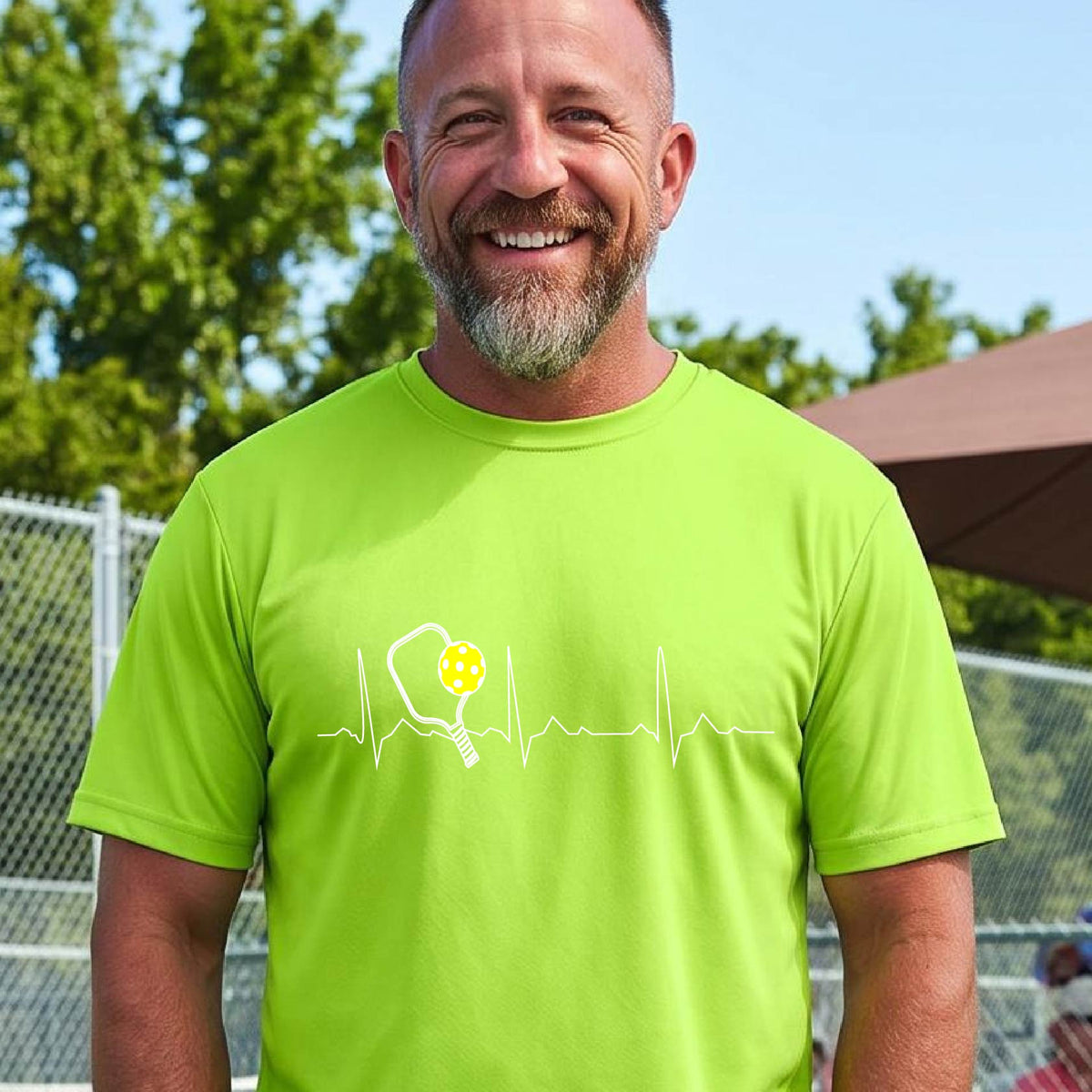 A close-up of a man in a lime green short-sleeve t-shirt sporting a white pickleball graphic of a heartbeat EKG including a pickleball paddle and a yellow pickleball across the check, in front of a pickleball court.